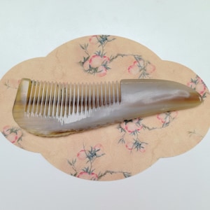 Puede incluir: Un peine de cuerno con dientes finos, que presenta un degradado natural de marrón claro a tonos translúcidos. El peine descansa sobre un papel color melocotón con un diseño floral, combinando funcionalidad y estética.