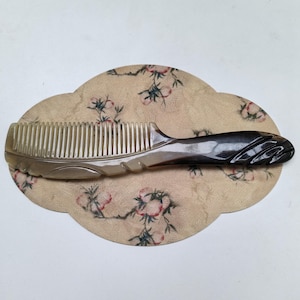 Puede incluir: Un peine para el cabello hecho de cuerno pulido, con un degradado de beige claro translúcido a gris carbón oscuro. El mango presenta ranuras diagonales talladas, mientras que los dientes son finos y están espaciados uniformemente.