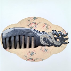 May include: A dark, polished horn comb featuring a carved dragon motif on the handle. The dragon body is detailed with white etched lines and a single small yellow stone eye. The comb has a series of fine, evenly spaced teeth along the edge.
