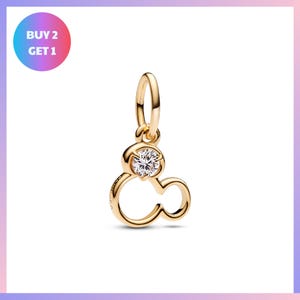 A gold-colored charm pendant with a clear gemstone in the center, resembling a stylized letter "M".