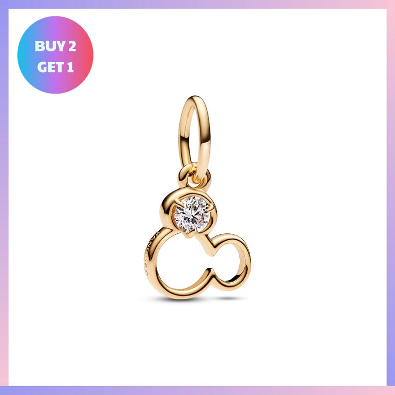 A gold-colored charm pendant with a clear gemstone in the center, resembling a stylized letter "M".