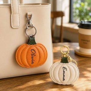 May include: Two pumpkin-shaped keychains with gold-tone metal hardware. One orange keychain features the name Olivia, and one cream-colored keychain features the name Emma. Both have green stems and are attached to a beige handbag and a wooden surface.