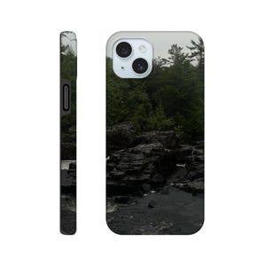 May include: A phone case featuring a scenic landscape of a river flowing through dark, rocky terrain, with lush green trees in the background under a cloudy sky. The case has a glossy finish and a camera cutout.