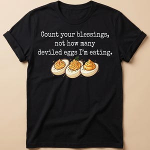 May include: A black short-sleeved t-shirt featuring the white text "Count your blessings, not how many deviled eggs I'm eating." Below the text are three illustrated deviled eggs with yellow yolks, garnished with green herbs and red spices.