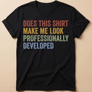 May include: A black short-sleeved t-shirt featuring the text "DOES THIS SHIRT MAKE ME LOOK PROFESSIONALLY DEVELOPED" printed in a stacked, multi-coloured design. The words are in red, yellow, green, and blue with a distressed, vintage-style texture.