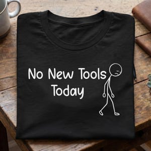 May include: A black short-sleeved t-shirt featuring white text that reads No New Tools Today, accompanied by a white line-art graphic of a sad stick figure walking with a slumped posture, printed on the front of the garment.