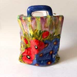 May include: A ceramic bag-shaped vase featuring a hand-painted design of vibrant red poppies and blue flowers against a green and purple striped background. The vessel has a glossy finish, a square base, and a single arched handle at the top.