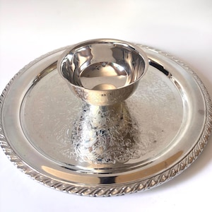 May include: A silver-toned metal serving set consisting of a circular tray and a central bowl. The tray features an intricate engraved floral pattern and a twisted rope-style border. The bowl is smooth, polished, and sits centered on the tray.