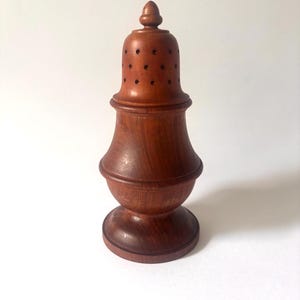 May include: A medium-brown wooden shaker with a smooth, turned finish. The object features a rounded base, a wider midsection, and a domed top with a small finial. Multiple small, circular holes are drilled into the upper dome section.