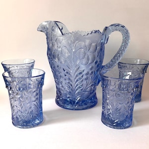 May include: A light blue glass pitcher and four matching tumblers, each featuring a detailed embossed floral pattern. The pitcher has a scalloped rim and a textured handle, while the tumblers have a straight cylindrical shape with matching relief.
