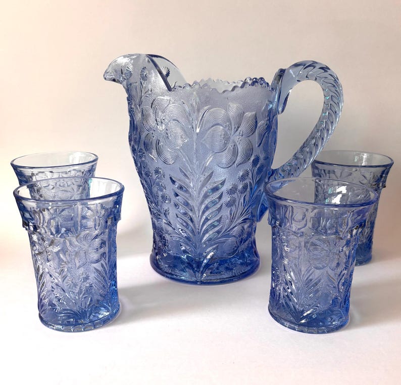 May include: A light blue glass pitcher and four matching tumblers, each featuring a detailed embossed floral pattern. The pitcher has a scalloped rim and a textured handle, while the tumblers have a straight cylindrical shape with matching relief.