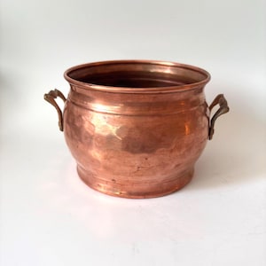 May include: A round, hammered copper pot featuring a wide, open top and two small, curved metal handles attached to the sides. The exterior surface displays a warm, metallic copper tone with visible indentations from the hammering process.