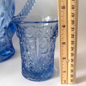 May include: A light blue glass tumbler with a raised floral pattern, positioned next to a wooden ruler showing a height of approximately 4.5 inches. A matching blue glass pitcher with a textured handle is partially visible in the background.