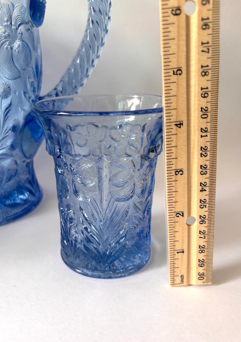 May include: A light blue glass tumbler with a raised floral pattern, positioned next to a wooden ruler showing a height of approximately 4.5 inches. A matching blue glass pitcher with a textured handle is partially visible in the background.