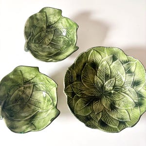 May include: Three green ceramic bowls arranged in a cluster, each featuring a detailed, embossed leaf pattern across the surface. The bowls vary in size, with a large bowl on the right and two smaller bowls on the left, all showcasing shades of green.