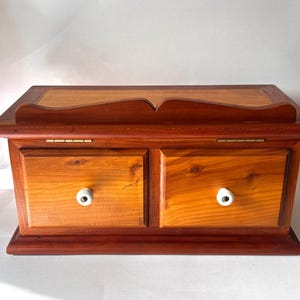 May include: A rectangular wooden chest featuring two front drawers with white ceramic knobs. The chest has a hinged lid with a decorative arched back panel. The wood displays a warm, reddish-brown grain pattern throughout the entire structure.