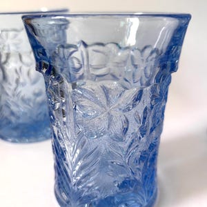 May include: A light blue glass tumbler featuring a raised, textured floral pattern across its exterior surface. The glass has a flared rim and a cylindrical body with a repeating botanical design of leaves and flowers molded into the transparent material.