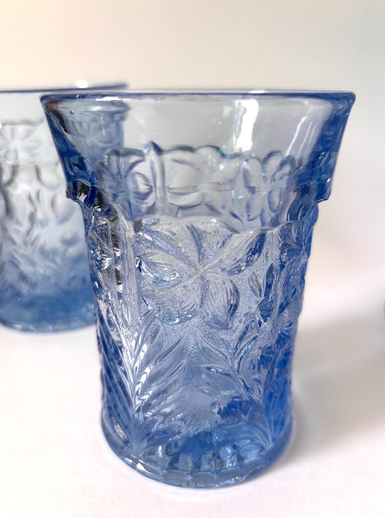 May include: A light blue glass tumbler featuring a raised, textured floral pattern across its exterior surface. The glass has a flared rim and a cylindrical body with a repeating botanical design of leaves and flowers molded into the transparent material.