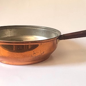 May include: A round copper skillet with a polished metallic exterior and a silver-toned interior lining. The pan features a long, straight, dark brown wooden handle attached to the side with a metal rivet, providing a contrast to the copper body.