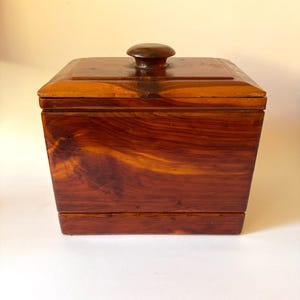 May include: A rectangular wooden box with a polished reddish-brown finish and visible wood grain patterns. The box features a flat lid with a centered, rounded knob handle. The construction shows a distinct base section and a smooth, glossy surface.