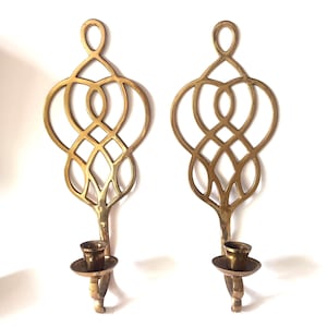 May include: A pair of matching brass wall sconces featuring an intricate, openwork lattice design at the top and a circular candle holder cup at the base. The metal has a polished, reflective gold-toned finish with a smooth surface.