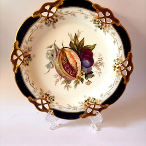 May include: A round porcelain plate featuring a central illustration of a sliced pomegranate and purple plums surrounded by green leaves. The plate has a wide navy blue border with gold-trimmed, openwork lattice edges and floral accents.