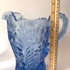 May include: A light blue pressed glass pitcher with a floral pattern and a scalloped rim, featuring a textured handle. A wooden ruler is placed vertically against the side, showing the pitcher stands approximately 8.5 inches in height.