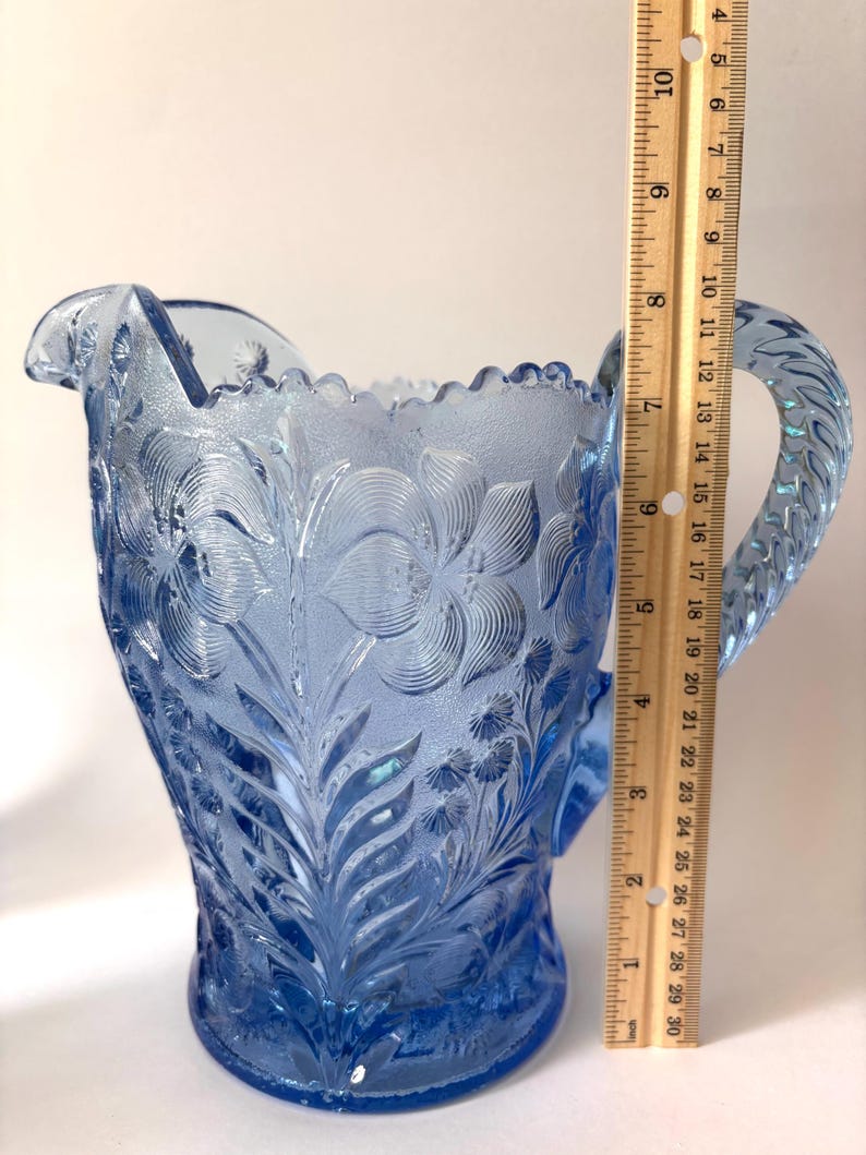 May include: A light blue pressed glass pitcher with a floral pattern and a scalloped rim, featuring a textured handle. A wooden ruler is placed vertically against the side, showing the pitcher stands approximately 8.5 inches in height.