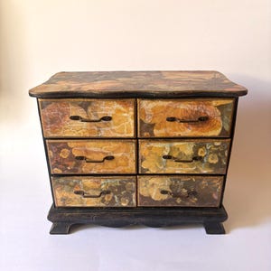 May include: A small wooden chest of drawers featuring six individual compartments. The exterior surfaces are decorated with a faded floral pattern in shades of yellow, orange, and brown. Each drawer is equipped with a thin, dark metal handle.