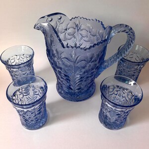 May include: A blue glass pitcher and four matching tumblers featuring a raised floral pattern. The pitcher has a scalloped rim and a textured handle, while each tumbler displays a consistent embossed botanical design across the exterior surface.