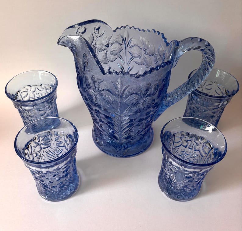 May include: A blue glass pitcher and four matching tumblers featuring a raised floral pattern. The pitcher has a scalloped rim and a textured handle, while each tumbler displays a consistent embossed botanical design across the exterior surface.