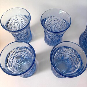 May include: Four light blue glass tumblers arranged in a square formation. Each tumbler features a textured exterior with a raised botanical pattern of leaves and ferns, and a sunburst design embossed on the interior base of the glass.