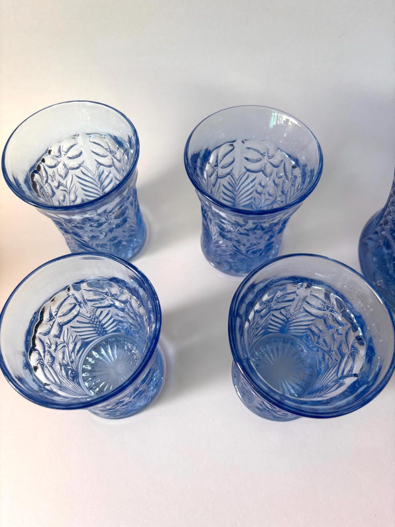 May include: Four light blue glass tumblers arranged in a square formation. Each tumbler features a textured exterior with a raised botanical pattern of leaves and ferns, and a sunburst design embossed on the interior base of the glass.