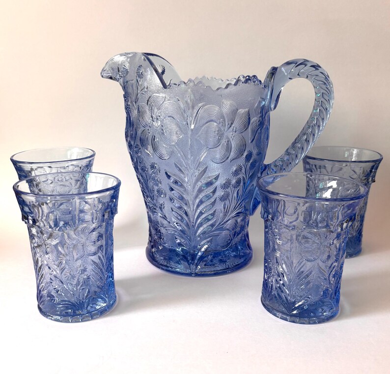 May include: A light blue glass pitcher and four matching tumblers, each featuring a detailed embossed floral pattern. The pitcher has a scalloped rim and a textured handle, while the tumblers have a straight cylindrical shape with matching relief.
