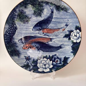 May include: A round decorative plate featuring a detailed illustration of four koi fish swimming among pine branches and white peony flowers. The artwork uses a palette of blue, white, and orange tones, with a thin gold-colored rim around the edge.