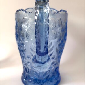 May include: A light blue glass pitcher featuring a raised floral pattern across its surface. The pitcher has a scalloped rim and a textured, vertical handle. The glass is translucent, showing a consistent blue hue throughout the entire piece.