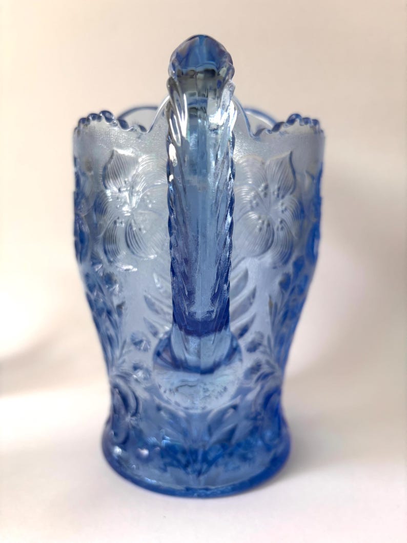 May include: A light blue glass pitcher featuring a raised floral pattern across its surface. The pitcher has a scalloped rim and a textured, vertical handle. The glass is translucent, showing a consistent blue hue throughout the entire piece.