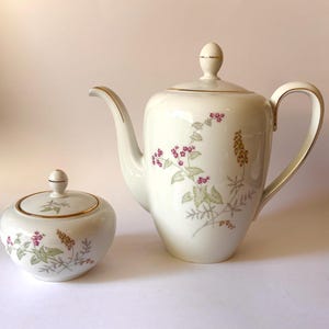 May include: A cream-colored porcelain coffee pot and a matching sugar bowl with a lid. Both pieces feature a delicate botanical pattern of small pink flowers, green leaves, and golden-brown stalks, accented with thin gold trim on the rims and lids.