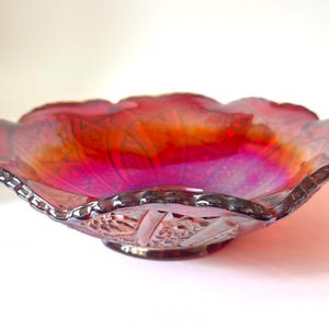 May include: A round, shallow glass bowl featuring a vibrant red and orange iridescent finish. The bowl has a scalloped, ruffled edge and a textured, pressed pattern on the exterior surface, including geometric and floral-like motifs.