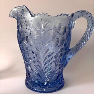 May include: A light blue glass pitcher featuring a textured surface with embossed floral and leaf patterns. The vessel has a scalloped rim, a curved pouring spout, and a twisted rope-style handle extending from the side of the body.