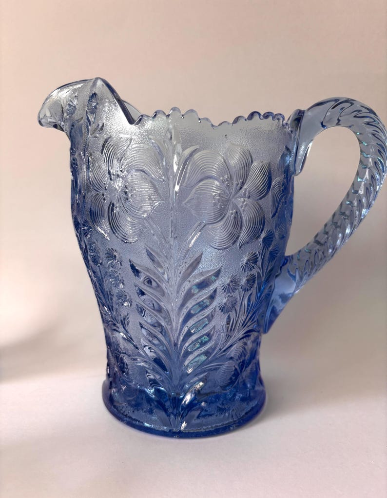 May include: A light blue glass pitcher featuring a textured surface with embossed floral and leaf patterns. The vessel has a scalloped rim, a curved pouring spout, and a twisted rope-style handle extending from the side of the body.