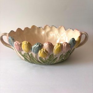 May include: A ceramic bowl with two handles featuring a raised, three-dimensional design of yellow, pink, and blue tulips with green leaves around the exterior. The interior of the bowl is a smooth, cream-colored glaze with a scalloped rim edge.