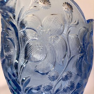 May include: A light blue glass pitcher featuring a raised, textured floral pattern. The surface displays intricate embossed details of large blossoms and smaller daisy-like flowers, with a translucent finish that catches the light across the glass.