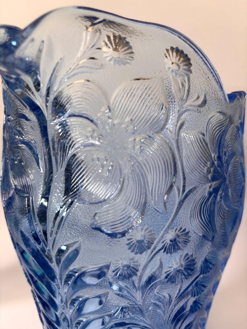 May include: A light blue glass pitcher featuring a raised, textured floral pattern. The surface displays intricate embossed details of large blossoms and smaller daisy-like flowers, with a translucent finish that catches the light across the glass.