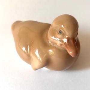 May include: A small, glossy ceramic figurine of a duckling in a seated position. The duckling is light brown with a slightly darker brown beak and a smooth, reflective finish. It has a rounded body, small wings tucked against its sides, and a detailed face.