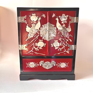 May include: A rectangular jewelry cabinet with a black frame and red doors and drawer. The front features inlaid mother-of-pearl peacock and floral designs, accented by ornate silver-toned metal hinges, a central circular latch, and a drawer pull.