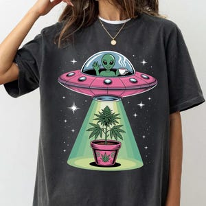 May include: Dark grey t-shirt with a pink UFO design, featuring a green alien waving and holding a cup. Below, a cannabis plant in a pink pot is lit by a green beam. The design includes stars.