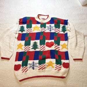 May include: A long-sleeve knit sweater featuring a cream base with a repeating grid pattern in red, blue, and green. The design includes embroidered motifs of yellow stars, green Christmas trees, white snowflakes, and red hearts throughout the front.