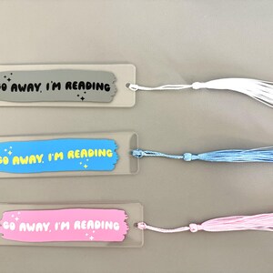 May include: Three rectangular bookmarks with rounded corners, each featuring a brushstroke design and the text "GO AWAY, I'M READING" with small star accents. The bookmarks come in gray, blue, and pink, each with a matching colored tassel attached.