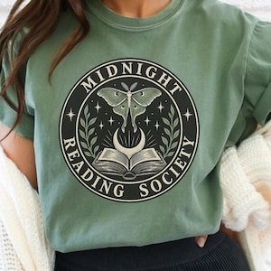May include: A moss green short-sleeve t-shirt featuring a circular graphic with a moth, an open book, and a crescent moon. The text reads MIDNIGHT READING SOCIETY. The shirt is worn with a white knit cardigan and black ribbed shorts.